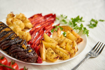 Fried potatoes, eggplant, bell pepper and cauliflower on white plate close up decorated with fork, fresh cherry tomatoes and green parsley