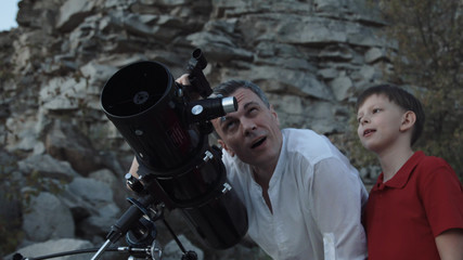 Father learning little son how to explore stars with telescope on rocky shore.
