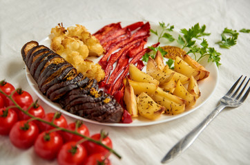 Fried potatoes, eggplant, bell pepper and cauliflower on white plate decorated with old silver fork, fresh red cherry tomatoes and green parsley