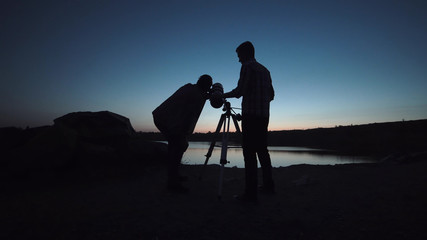 Silhouettes of people looking through telescope on shore of lake in dark.