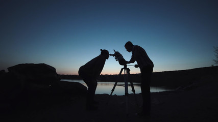 Silhouettes of people looking through telescope on shore of lake in dark.