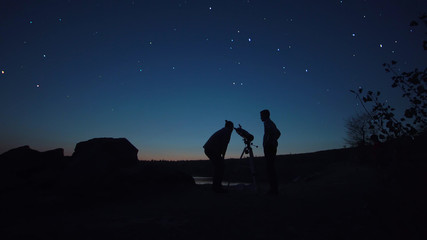 Black silhouettes of people looking through telescope on stars in dark twilight on shore.