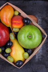 Red apple, dark blue grapes, green apple, yellow pear, green grapes, orange peach in a wooden box on a metallic background in retro style