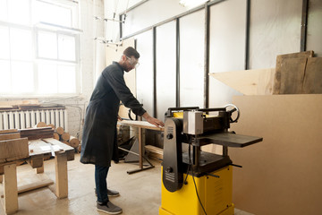 Young male carpenter using power tool for his work in small woodshop, working with stationary planer to reduce wooden boards thickness and smooth plank, cabinetmaker or joiner activity and woodworking