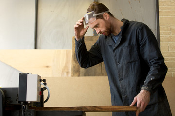 Worker in protective clothing puts massive wooden board in woodworking machine. Carpenter working on sawmill or lumber manufacture at machine tool. Small business and local entrepreneurship concept