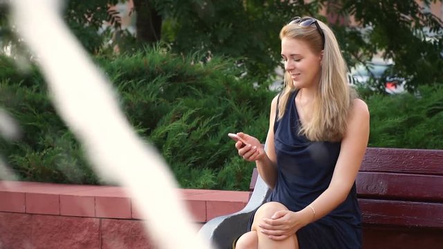 Happy girl using a smartphone in a city park sitting on a bench next to the fountain