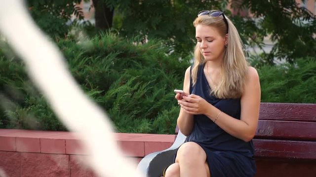 Happy girl using a smartphone in a city park sitting on a bench next to the fountain