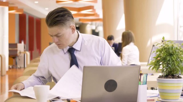 young nervous man feels stress and pressure because of difficulties and failure during working day with papers sitting at table with laptop PC in co working colorful business office space