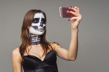 beautiful young of brown-haired woman with terrifying halloween skeleton makeup using pink smart phone on a gray background