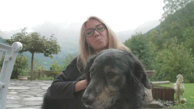 Affectionate young blonde woman in glasses embracing and petting cute black dog on cloudy autumn day outdoors in a park. Attractive girl stroking her pet on the ears and head.