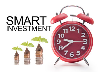 Business and financial concept.Little green plant on the stacks of coins with red twin bell alarm clock isolated over white background with text Smart Investment.