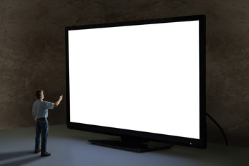 Man pointing tv remote control at world’s biggest television that towers over him shining bright light in a dimly lit room with clean blank screen for your message and image sending a powerful message