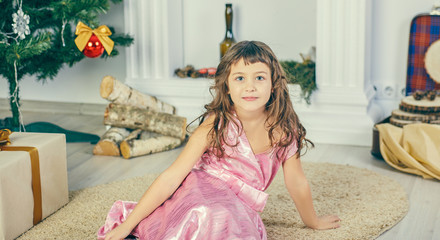 Obraz premium Cheerful cute little child girl with present. Kid is sitting near a gift box and Christmas tree indoors.