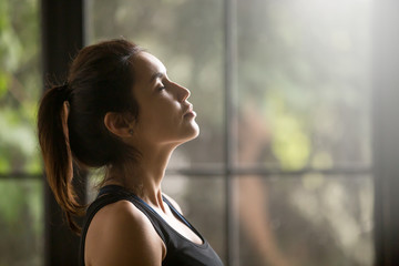 Profile portrait of young attractive yogi woman breathing fresh air, her eyes closed, meditation pose, relaxation exercise, working out wearing black sportswear top, close up image, window background