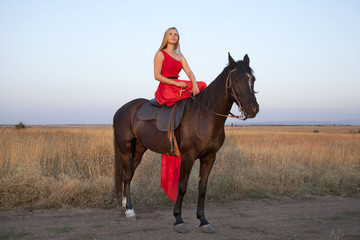 Slim blonde in a long red dress on horseback. Young woman looking into the distance enjoying an evening walk