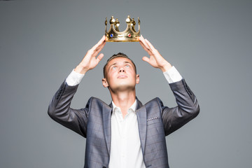 Young attractive man in a suit holding above his head a golden crown on a gray background