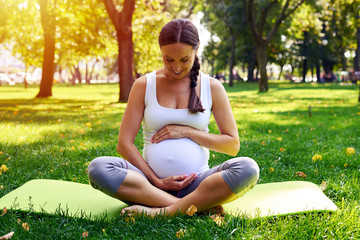 Fototapeta premium Lovely pregnant woman embracing belly on yoga mat in park