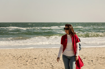 Young woman (brunette) in a red cardigan and light blue jeans, with a backpack, walks along the beach.