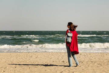 Young woman (brunette) in a red cardigan and light blue jeans, with a backpack, walks along the beach.