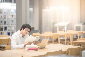 Young Asian man university student smiling and reading book in library, education research and self learning in college life concepts