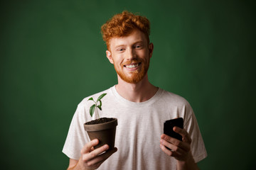 Fototapeta premium Portrait of young smiling redhead bearded young man, holding spotted plant and mobile phone