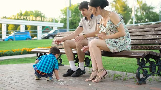 Young parents posing on bench and drawing on asphalt with son. Young man and woman sitting on bench and helping little son to draw with chalk on ground.