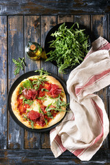 Whole homemade pizza with cheese and bresaola, served on black plate with fresh arugula, olive oil and kitchen towel over old wooden plank background. Flat lay.