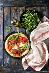 Poster de jardin Pizzeria Whole homemade pizza with cheese and bresaola, served on black plate with fresh arugula, olive oil and kitchen towel over old wooden plank background. Flat lay.