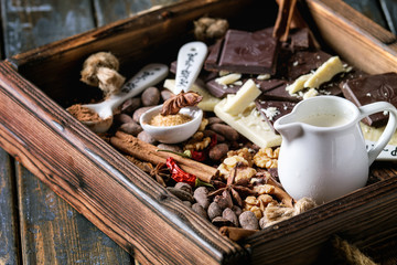 Ingredients for cooking hot chocolate. White and dark chopping chocolate, cocoa powder, cocoa beans, cream, cinnamon, sugar in spoons in wood tray over old wooden background. Close up