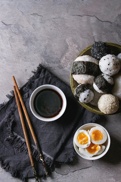 Bowl with different size rice balls with black sesame and seaweed nori, served with soft boiled eggs, soy sauce, chopsticks over gray table. Asian style dinner. Top view with space