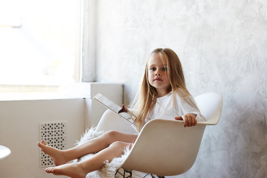 Picture of cute little girl familiar with gadgets sitting barefooted on comfortable armchair by the window, holding digital tablet, using free wifi, watching video online on how to bake cookies
