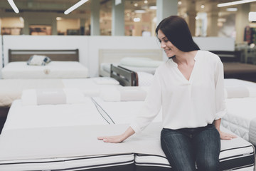 A woman chooses a mattress in a store. She sits on it and examines it. - obrazy, fototapety, plakaty