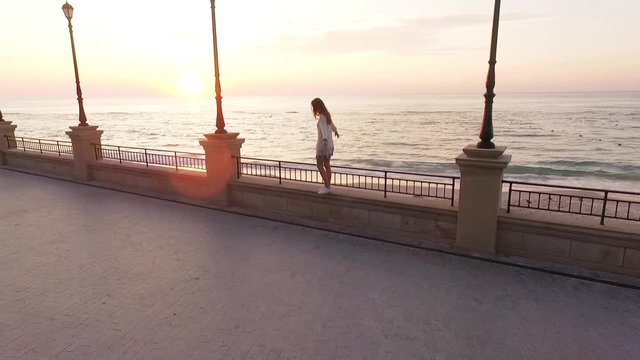slomo from drone young woman with long hair in white outfit walking along parapet of embankment balancing backside with empty beach background on sunrise