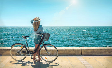 Blonde woman in summer hat with her bicycle walking coastline by