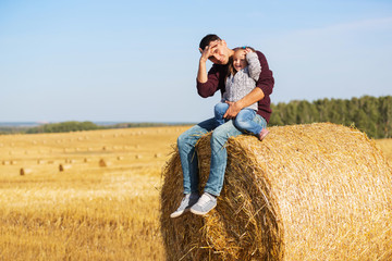 Happy father and 2 year old girl sitting on hay bale in harvested field