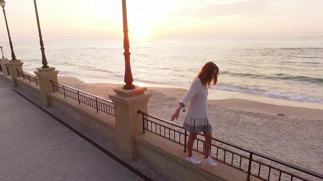 aerial footage skinny long-haired girl walking along parapet of embankment keeping balance looking far into sea on sunset in slow motion