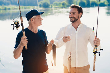 The old man and his adult son are standing on the river bank and posing with the fish that they just caught