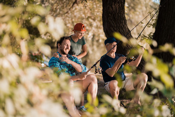 Fototapeta premium View from behind the trees. The man, his little son and his elderly father are fishing for spinning on the river bank