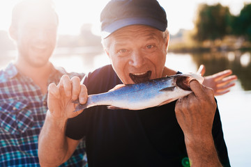 An old man in a black T-shirt posing while holding a fish. He pretends that he wants to eat her