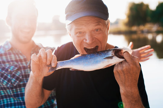 An old man in a black T-shirt posing while holding a fish. He pretends that he wants to eat her