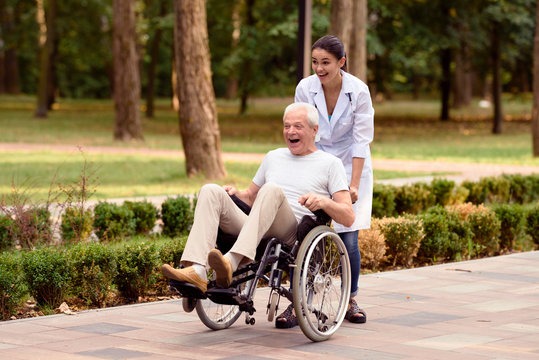 The doctor rolls an elderly patient on a wheelchair in the park. They are having fun