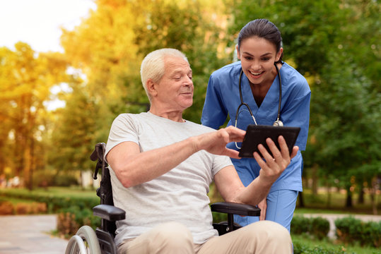 An old man in a wheelchair proudly shows a happy nurse something in his tablet