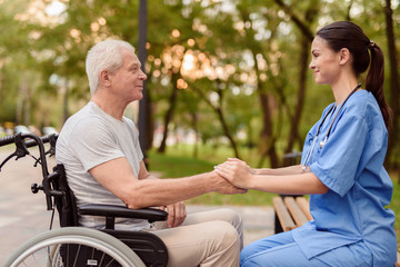 Fototapeta premium An old man who sits in a wheelchair and a nurse who sits next to the bench holding hands