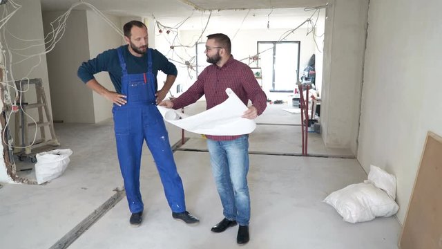 Male architect with blueprints talking to his worker about project at new home
