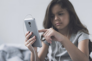 A little black-haired girl in white headphones with a gray smartphone in her hands switches music