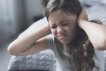 The black-haired girl sits next to the bed frowning and covering her ears with her hands