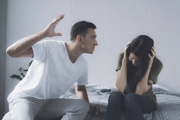 Fototapeta premium Family problems. A man swings to hit a woman who sits next to him on the bed and covers herself with her hands