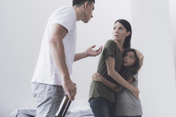 Evil drunken man with a bottle in his hands screams at his wife and daughter who are facing him embracing in fear