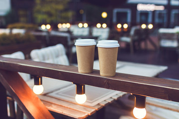 Two cups takeaway coffee on cafe veranda with bulb lights. Romantic evening