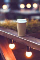 Cup takeaway coffee on cafe veranda with bulb lights. Coffee to go. 